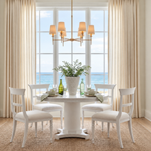 Load image into Gallery viewer, A bright dining room with a round white pedestal table, four white upholstered chairs, a brass chandelier, beige curtains, and a large window overlooking the ocean.
